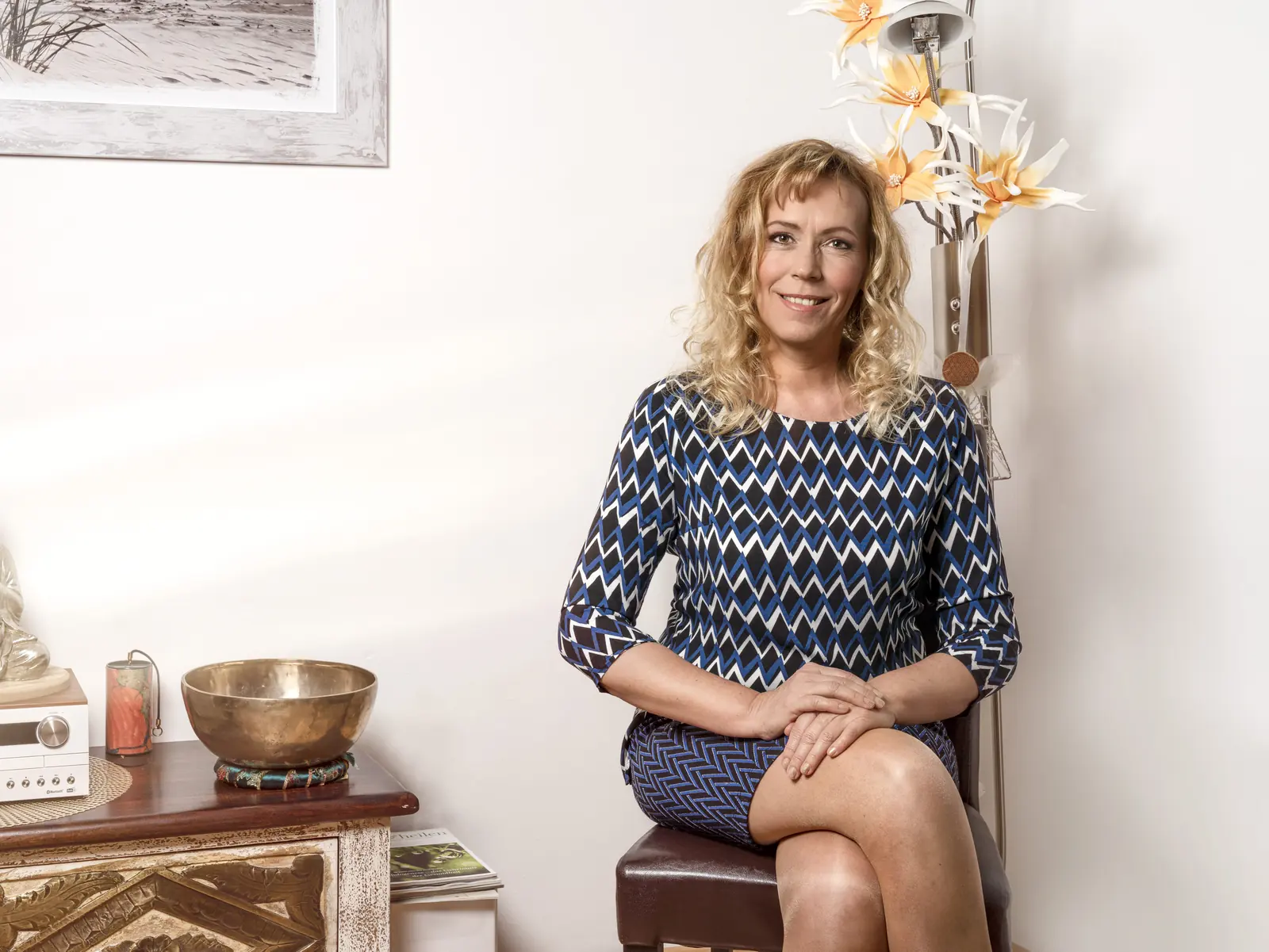 Bettina Ziperzik sitzt auf einem Hocker in ihrer Praxis, daneben eine Kommode mit Buddha und Klangschale